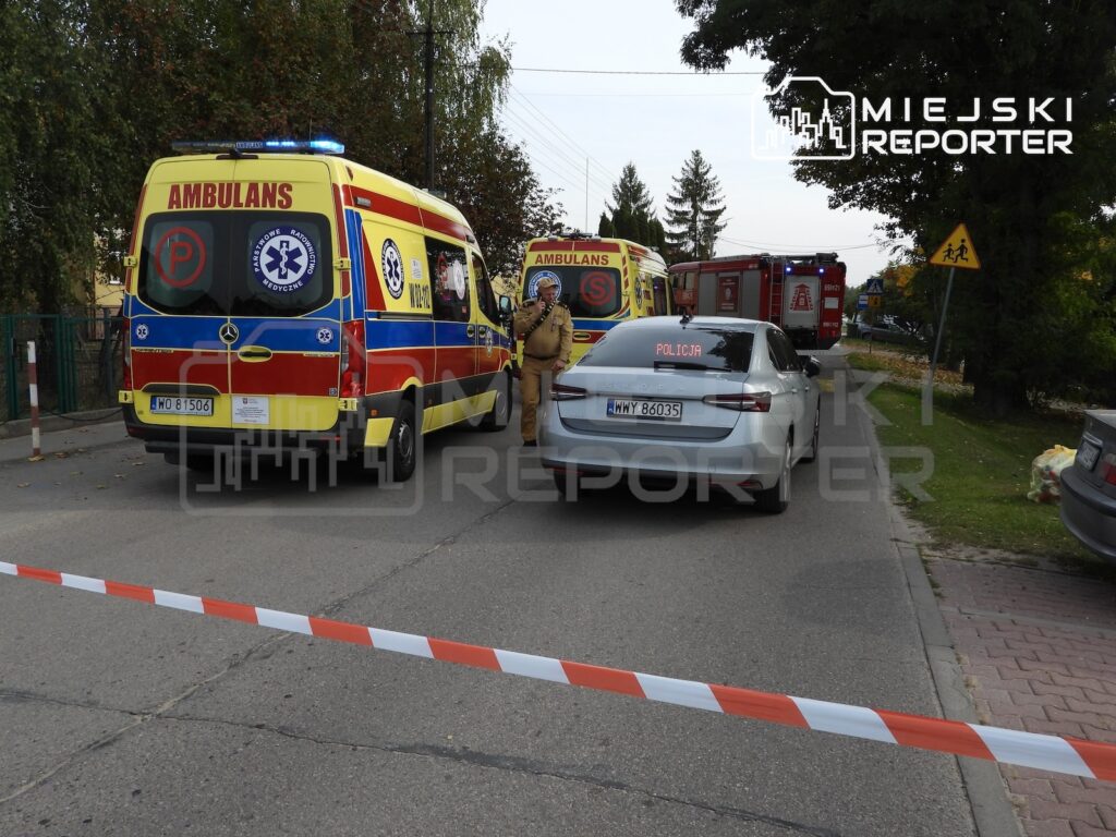 Ambulanse, radiowóz policji i wóz strażacki na zamkniętej taśmą ulicy, przy której stoją służby ratunkowe.