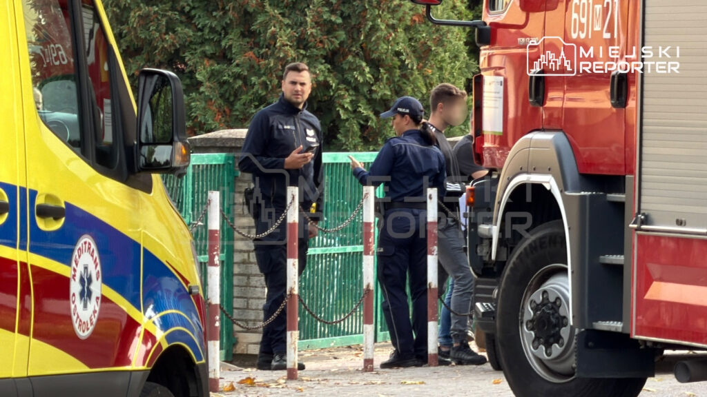 Ratownicy medyczni i strażacy rozmawiają przy karetce i wozie strażackim obok zielonej bramy szkolnej