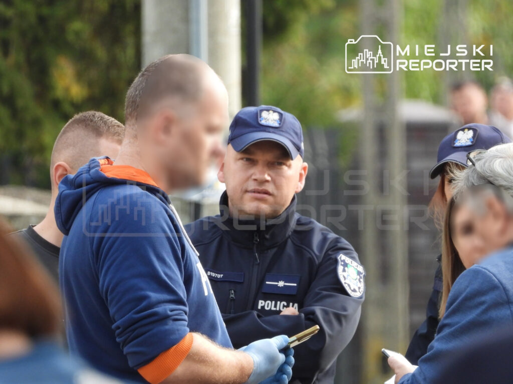 Policjant rozmawia z mężczyzną w rękawiczkach, trzymającym telefon, na zewnątrz obok innych osób.