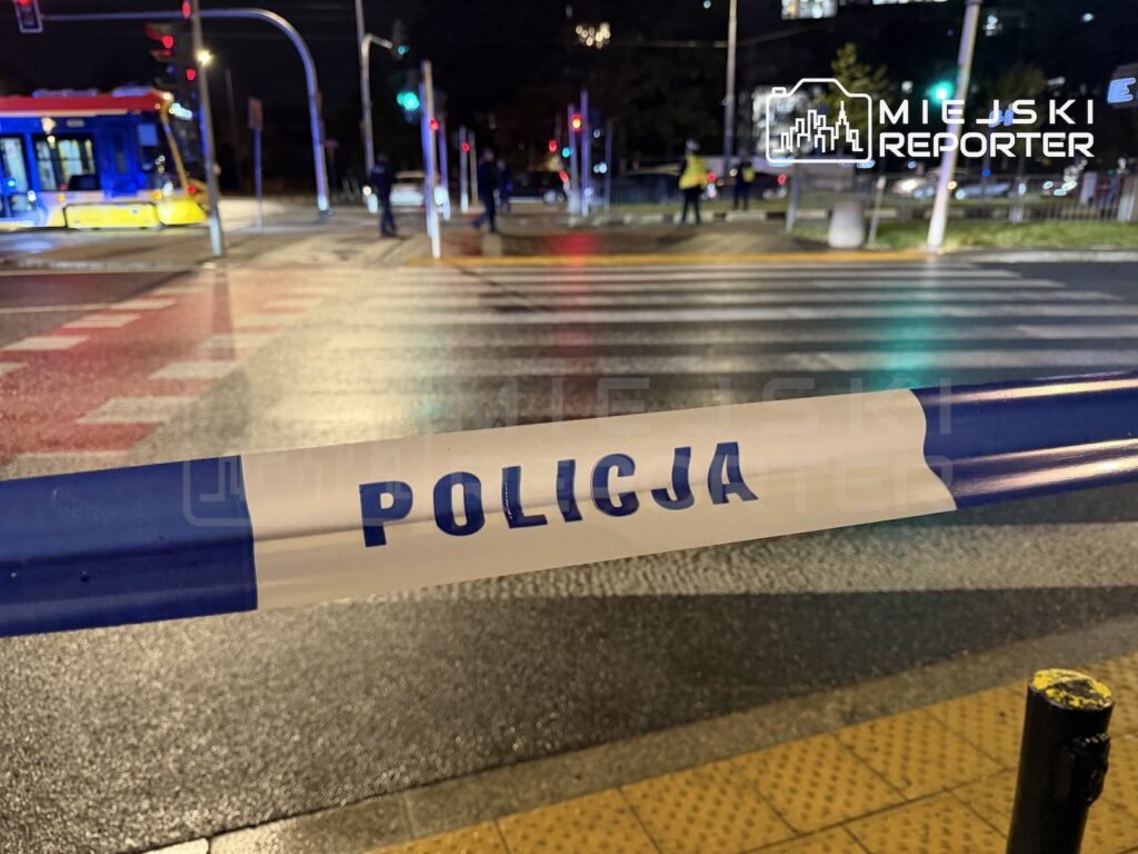 Taśma z napisem „POLICJA” odgradza nocą przejście dla pieszych; w tle widać tramwaj i skrzyżowanie.