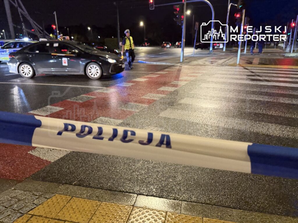Taśma policyjna z napisem „POLICJA” odgradza nocne skrzyżowanie z przejściem dla pieszych i samochodem.