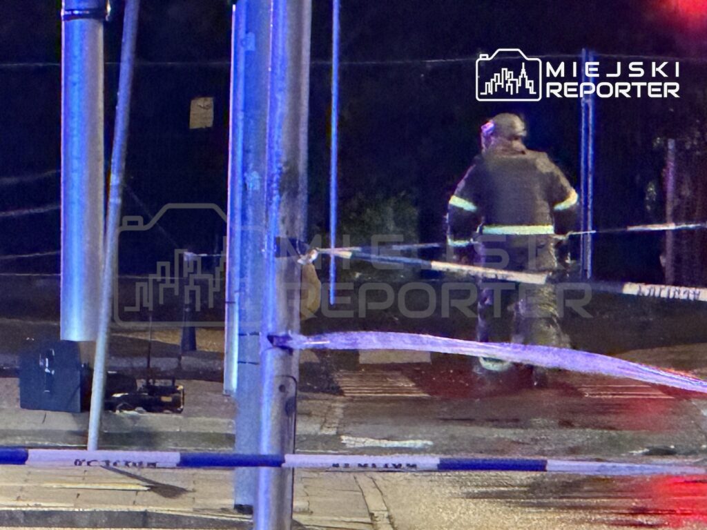 Strażak w odblaskowym ubraniu stoi za policyjną taśmą na nocnym skrzyżowaniu oświetlonym niebieskimi światłami.