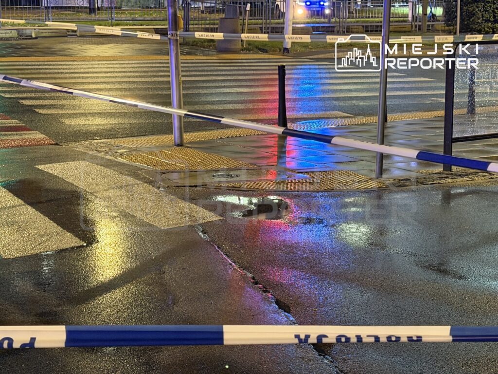 Policyjny radiowóz z włączonymi niebieskimi światłami za taśmą zabezpieczającą mokre przejście dla pieszych nocą.
