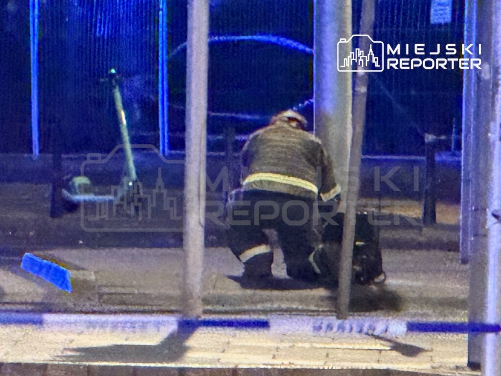Osoba w ciemnym ubraniu z odblaskowymi pasami pochyla się nad leżącym przedmiotem przy przeszklonej wiacie przystankowej.