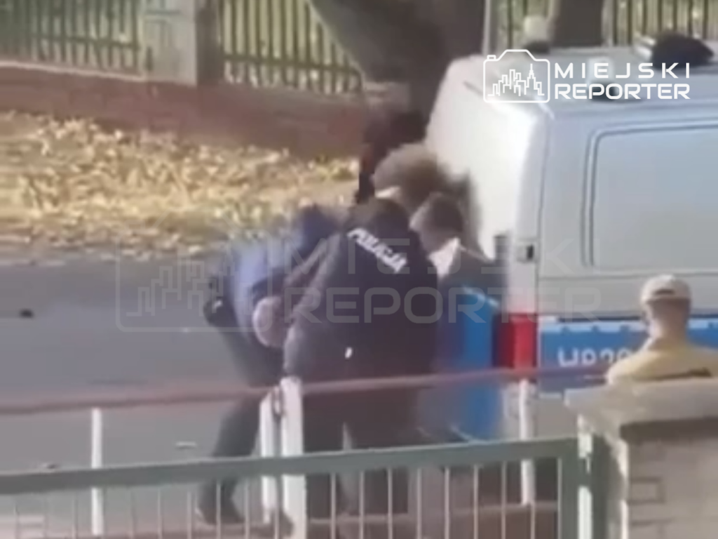 Policjanci obezwładniają mężczyznę przy zaparkowanym radiowozie, za metalowym ogrodzeniem.