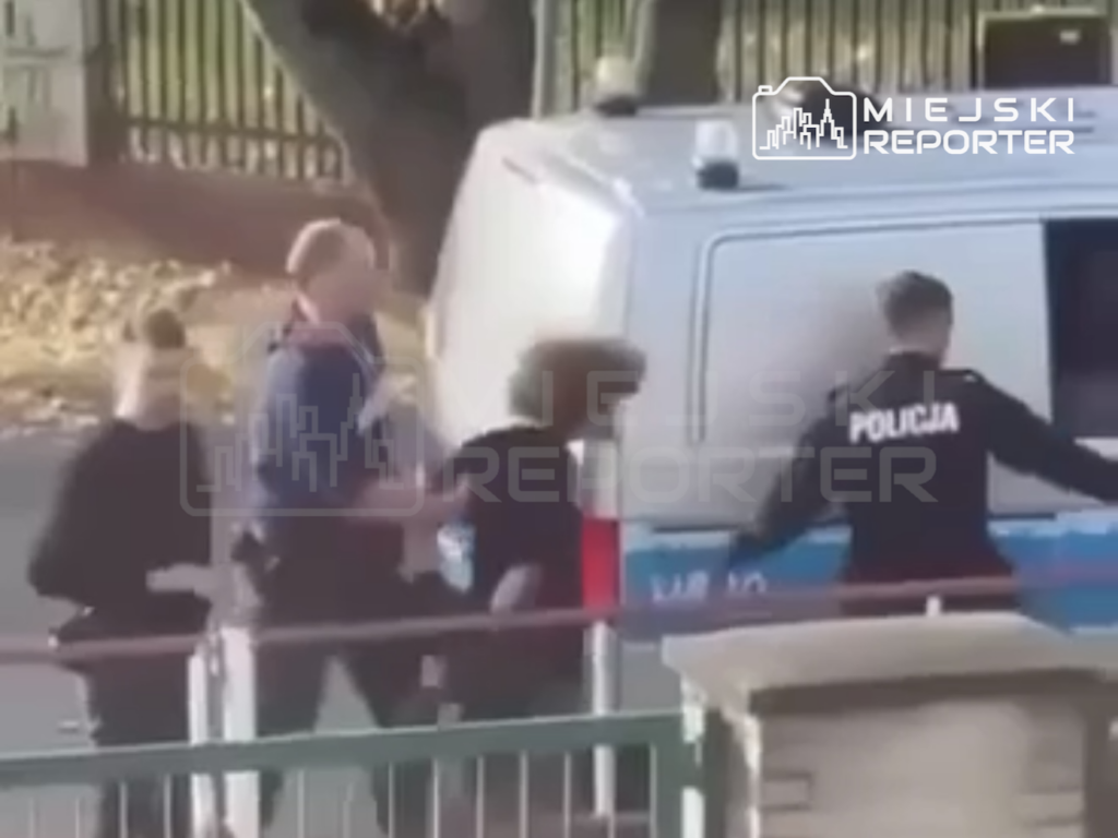 Policjant prowadzi osobę w ciemnym ubraniu obok policyjnego furgonu, za barierką uliczną.