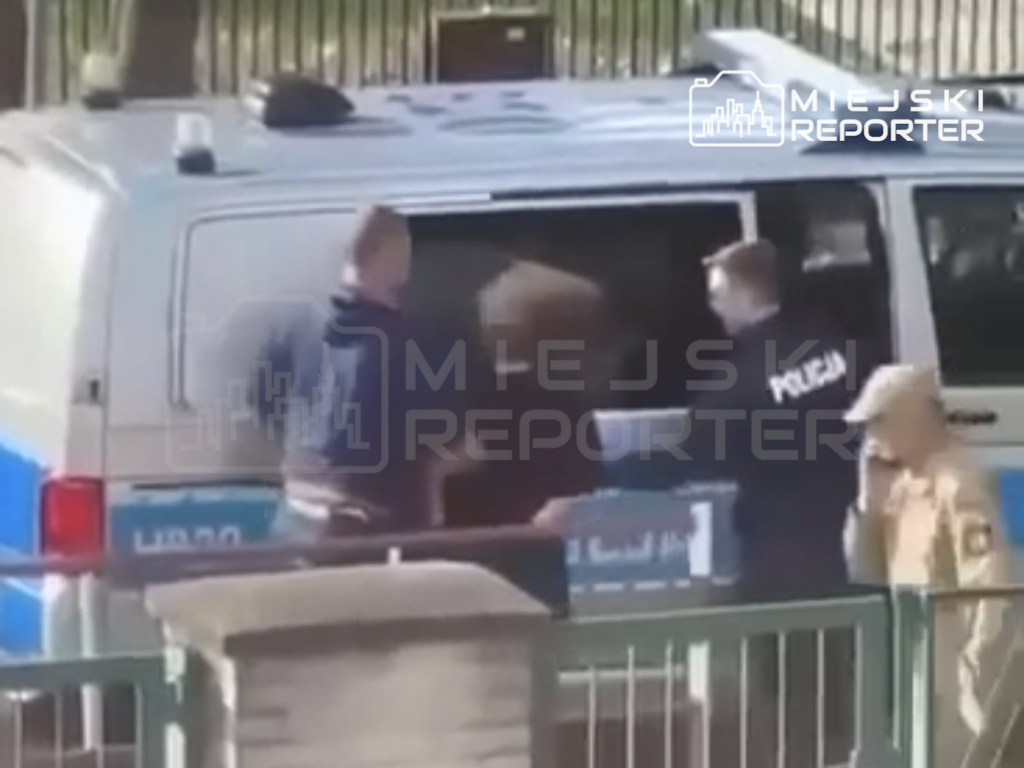 Policjanci stoją przy otwartych drzwiach policyjnego vana, za zielonym ogrodzeniem budynku.