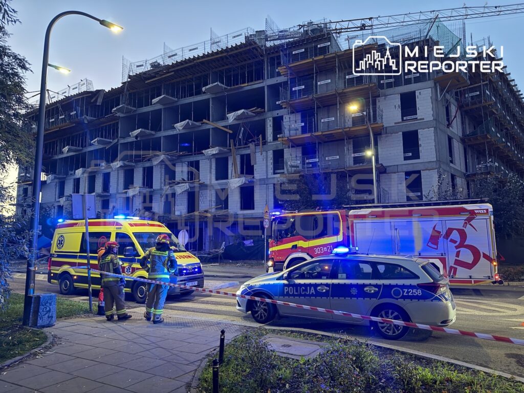 Policjanci i ratownicy przy karetkach przed budynkiem w budowie, zabezpieczonym taśmą policyjną o zmierzchu.