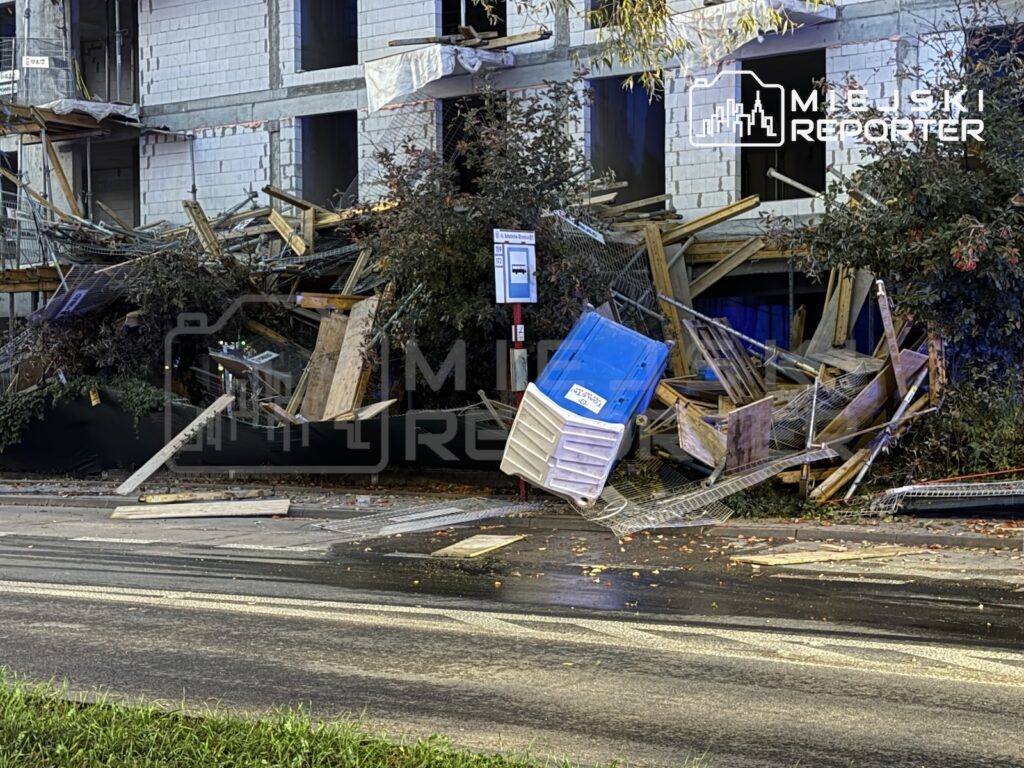 Zawalona drewniana konstrukcja przy budynku mieszkalnym, przewrócony niebiesko-biały pojemnik i gruz na chodniku przy ulicy.