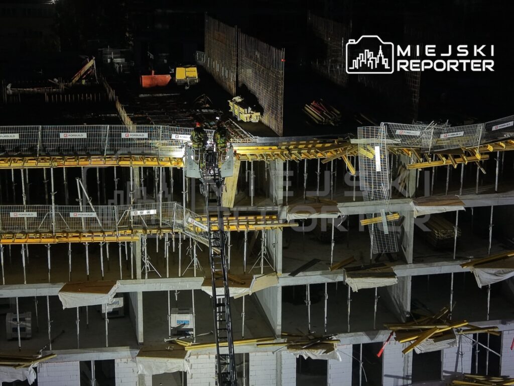 Nocny plac budowy z betonową konstrukcją budynku, rusztowaniami i ogrodzeniami zabezpieczającymi teren.