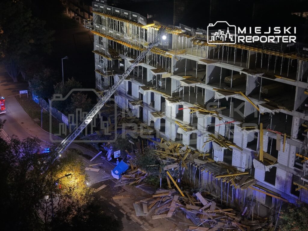 Nocny widok na uszkodzony, częściowo zawalony budynek mieszkalny i gruz na placu budowy, oświetlone reflektorami.