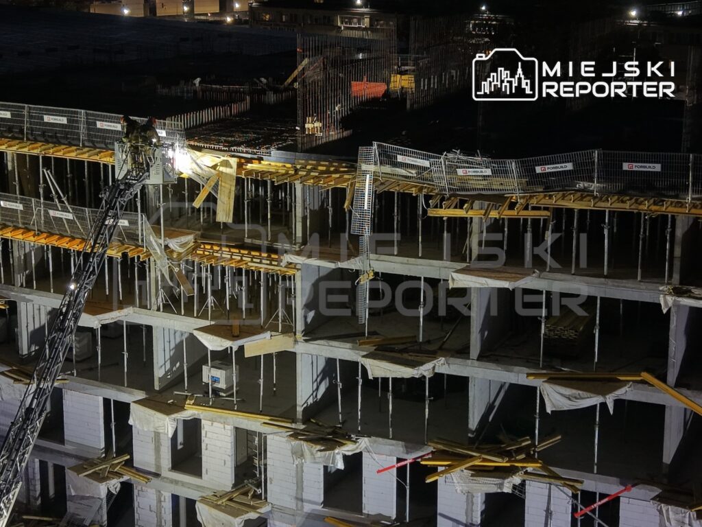 Nocny widok placu budowy z betonową konstrukcją budynku, rusztowaniami i barierkami zabezpieczającymi