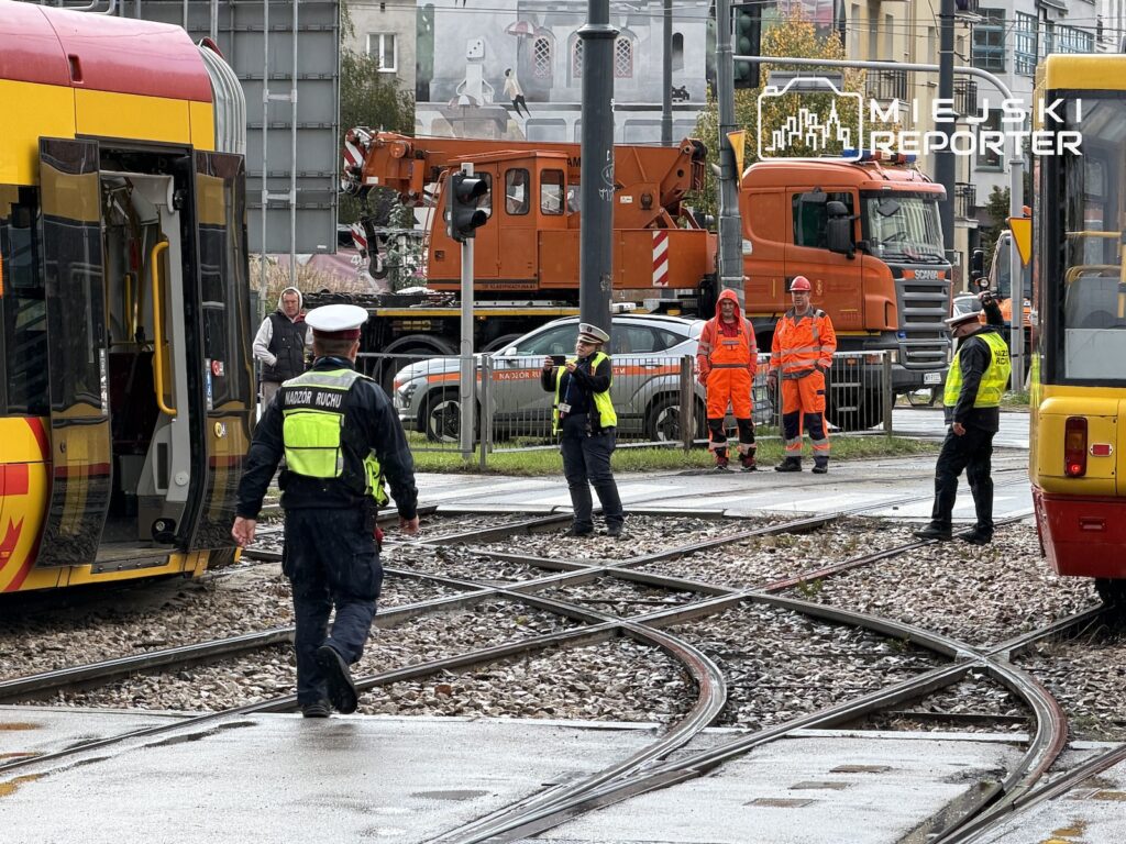 Pracownicy służb i kolejarze stoją przy torowisku między tramwajami, obok dźwigu i samochodu technicznego.