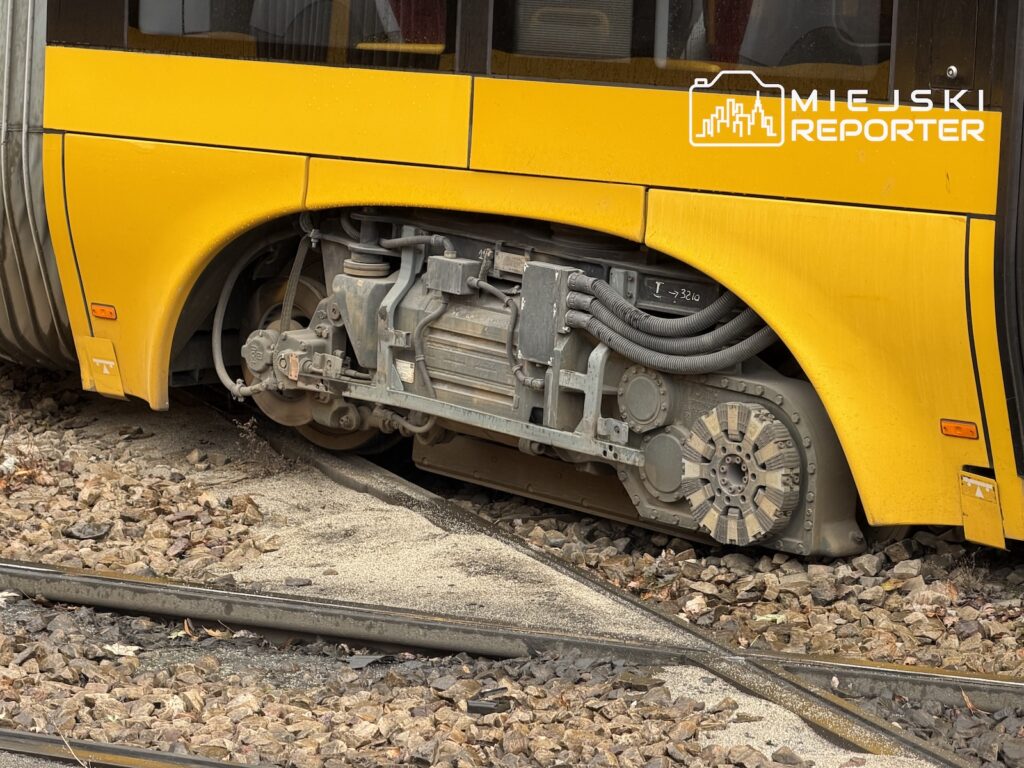 Widok podwozia żółtego tramwaju z odsłoniętymi elementami, stojącego na torach zasypanych kamieniami.