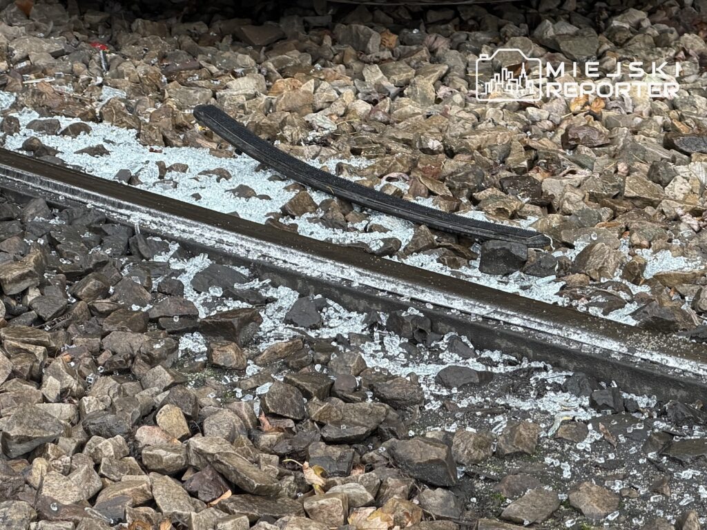 Uszkodzony fragment torowiska tramwajowego z odgiętą szyną, otoczony kamiennym tłuczniem.