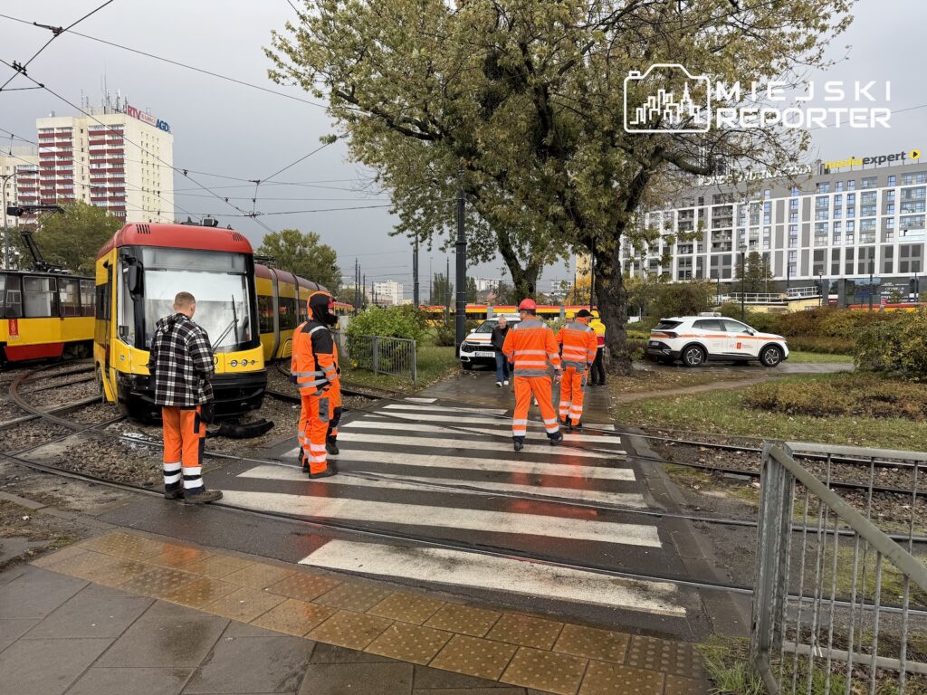 Pracownicy w pomarańczowych strojach stoją przy czerwono-żółtym tramwaju na przejściu przez tory.