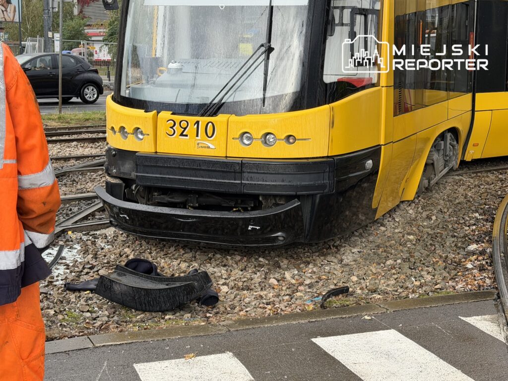 Żółty tramwaj z uszkodzonym przednim zderzakiem stoi na torach; obok leży oderwany element, a pracownik w odblaskowym stroju