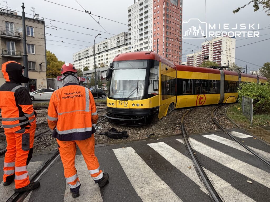 Żółto-czerwony tramwaj stoi poza torami przy przejściu dla pieszych; obok dwóch pracowników w odblaskowych uniformach.