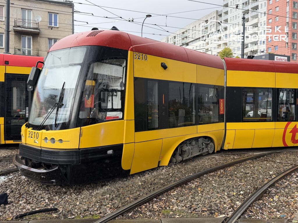 Czerwono-żółty tramwaj stoi na torach przy ulicy, obok drugiego składu i bloków mieszkalnych.