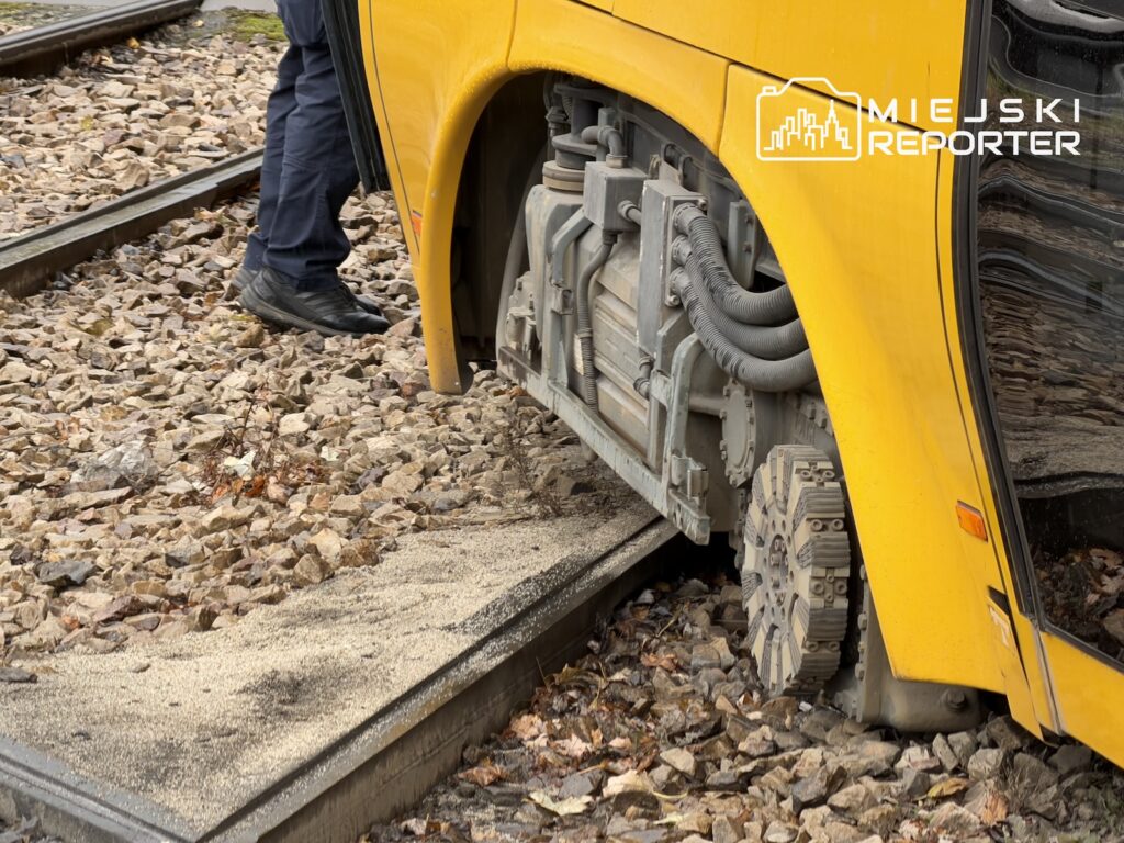 Koło żółtego tramwaju znajduje się poza szyną, obok torowiska i kamienistego podłoża.