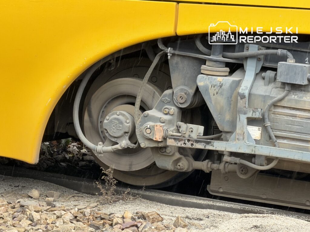 Koło żółtego tramwaju i jego podwozie nad torowiskiem z tłuczniem, widoczne z bliska