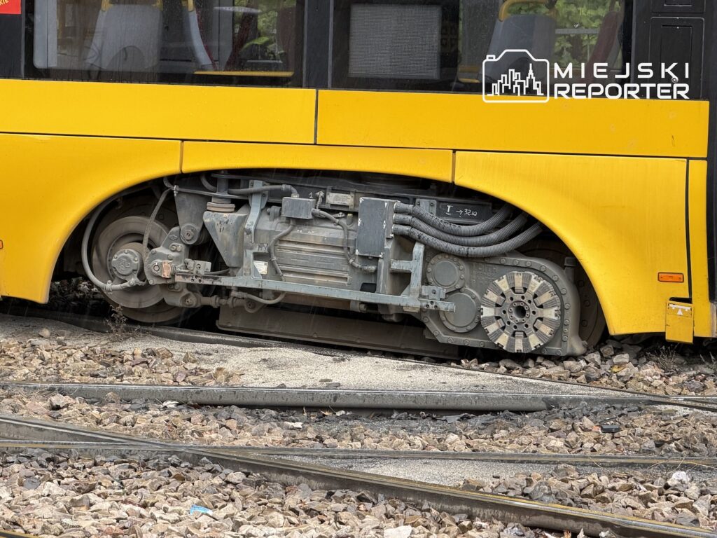 Odsłonięte podwozie żółtego tramwaju z kołem i mechanizmami widocznymi nad torowiskiem.
