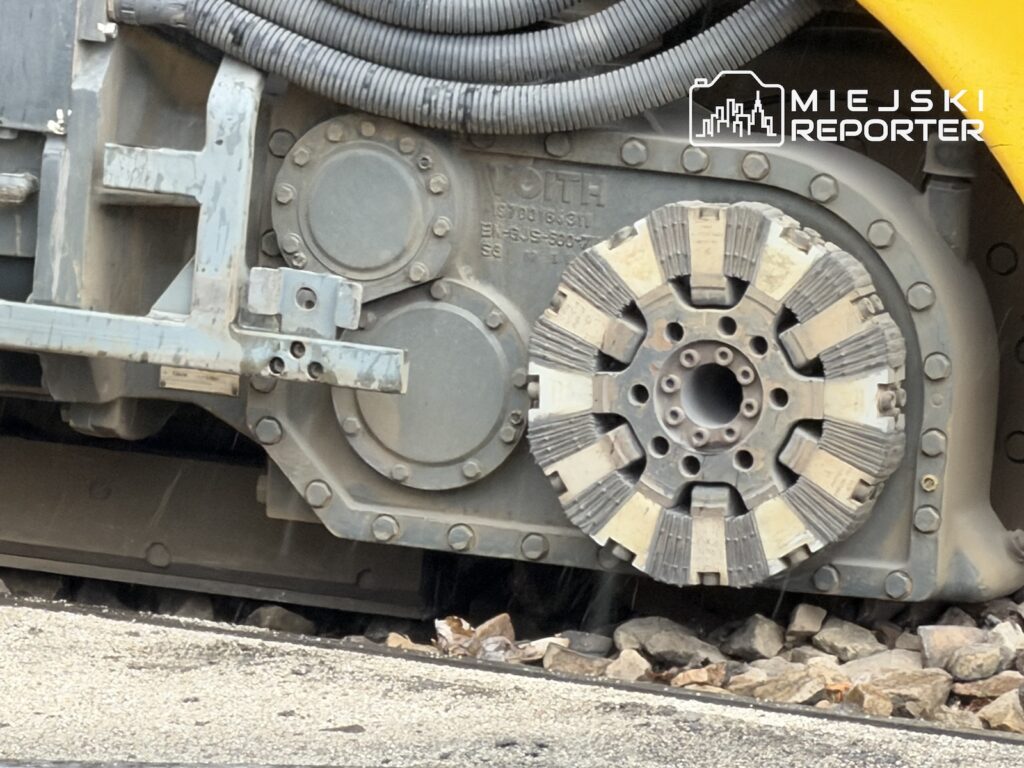 Zbliżenie na odsłonięty mechanizm podwozia tramwaju, tuż nad torowiskiem i kamieniami.