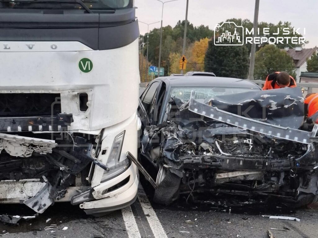 Rozbity przód samochodu osobowego obok uszkodzonej ciężarówki na jezdni, przy wraku stoją ratownicy w odblaskowych ubraniach.