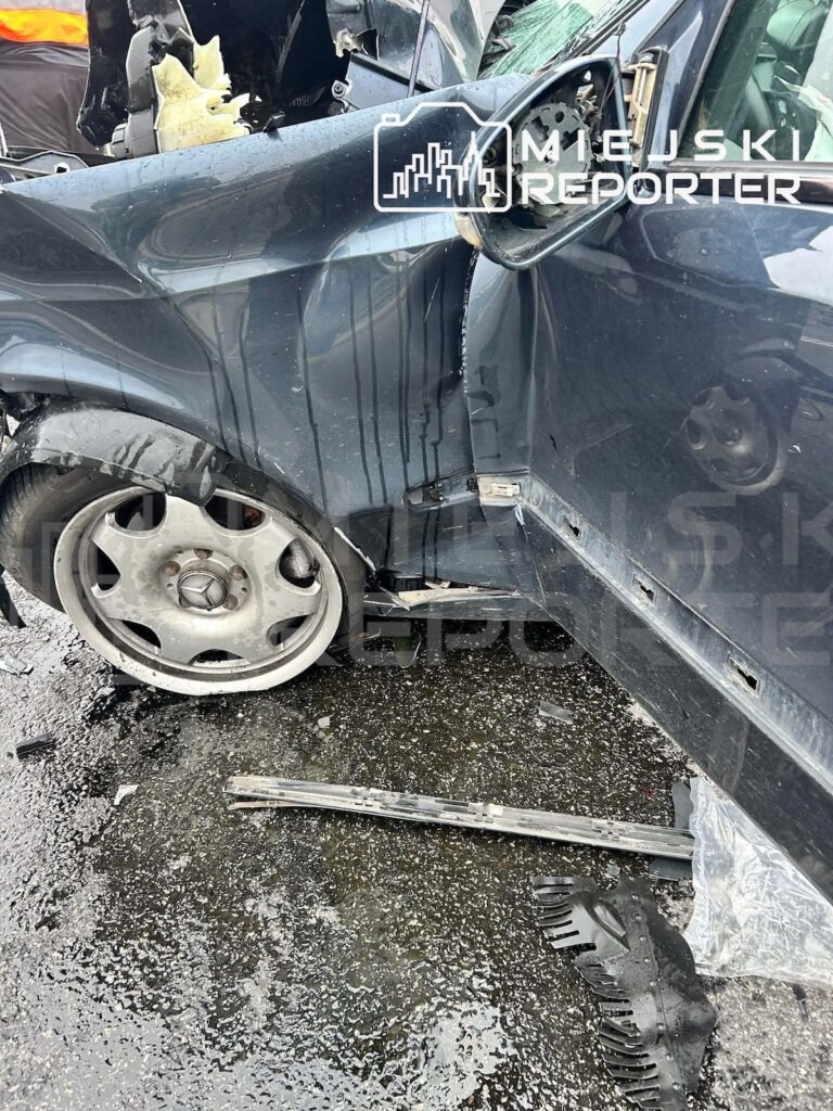 Rozbity ciemny samochód z wgniecionym bokiem i rozbitymi szybami, na jezdni leżą odłamki i płyny.