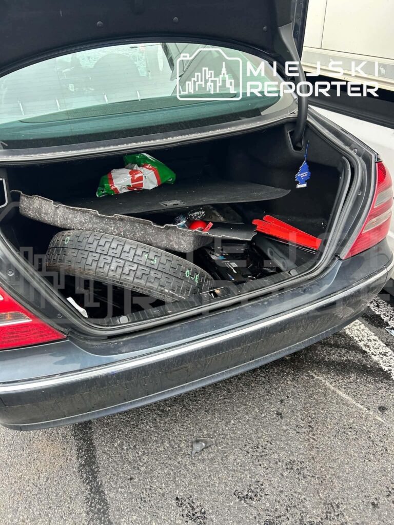 Otwarty bagażnik ciemnego samochodu z uszkodzoną osłoną i kołem zapasowym na podłodze parkingu.