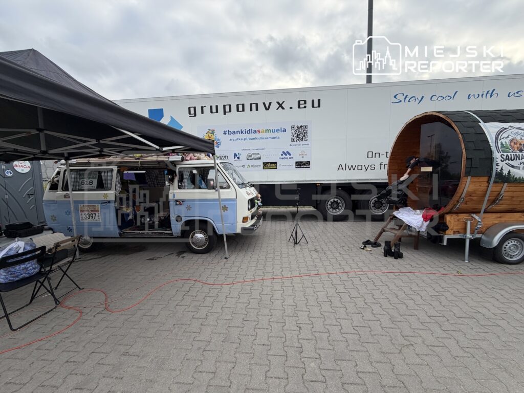 Biały samochód dostawczy pod namiotem i drewniana beczka na przyczepie stoją przed ciężarówką z napisem „gruponvx.eu”.