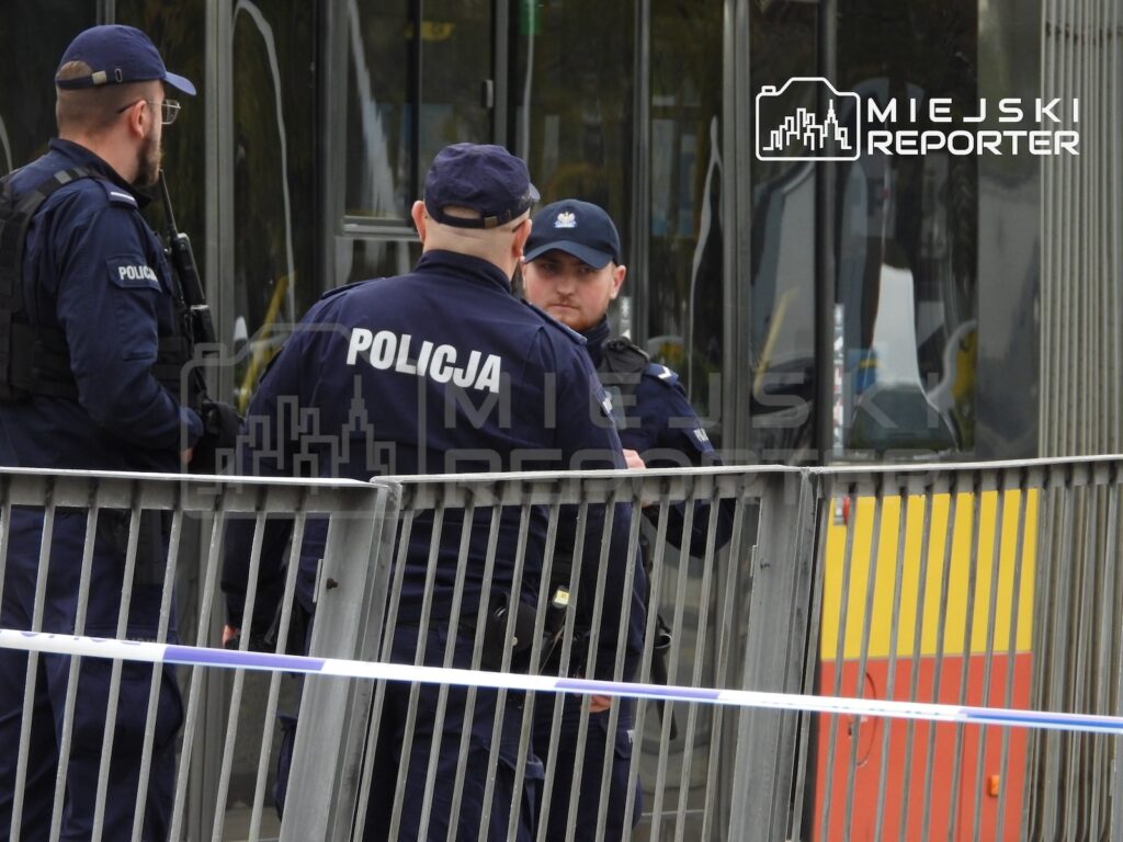 Policjanci w mundurach rozmawiają przy barierkach i taśmie policyjnej, obok przeszklonego budynku.