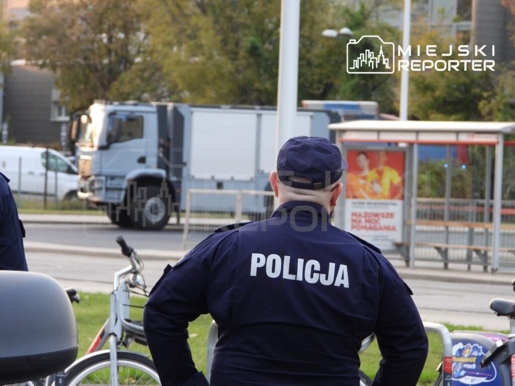 Policjant w granatowej kurtce z napisem „POLICJA” stoi tyłem na ulicy, obok stojaków rowerowych.