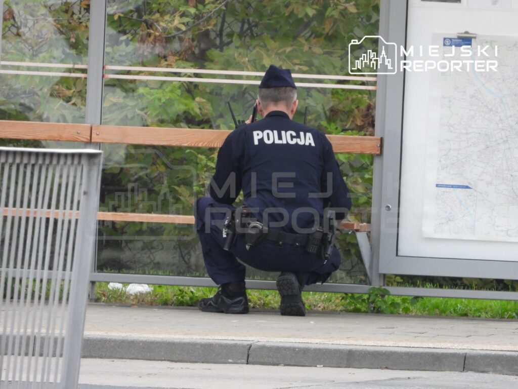 Policjant w mundurze i czapce kuca przy przystankowej wiacie, obok tablicy z mapą.