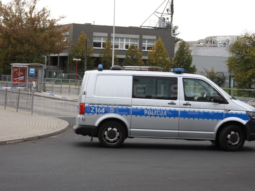Policyjny radiowóz stoi przy ogrodzonym terenie przed budynkiem, obok przystanku autobusowego.