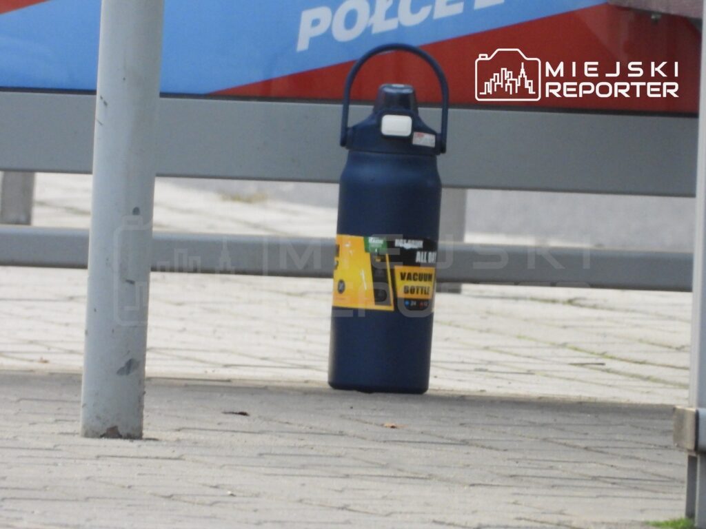 Ciemny pojemnik z żółtą etykietą stoi na chodniku przy metalowej barierce.