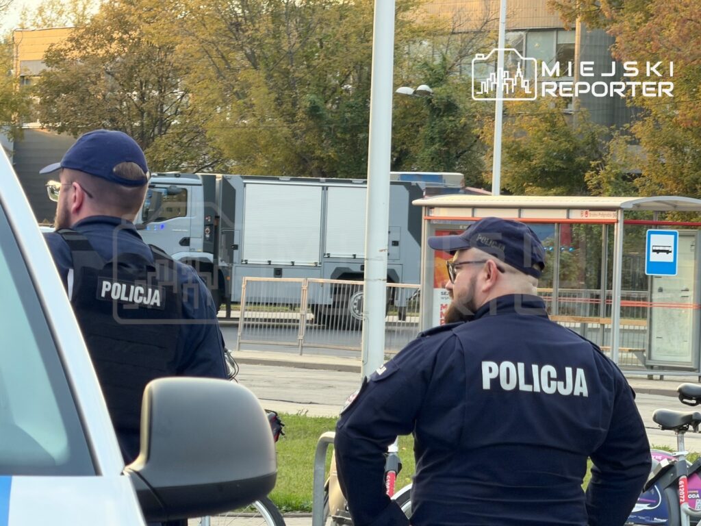 Dwóch policjantów w granatowych mundurach stoi przy radiowozie obok przeszklonej wiaty przystankowej.
