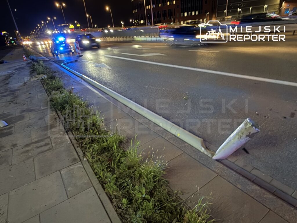 Przewrócona latarnia leży na chodniku przy nocnej ulicy, w tle stoi radiowóz z włączonymi niebieskimi światłami.