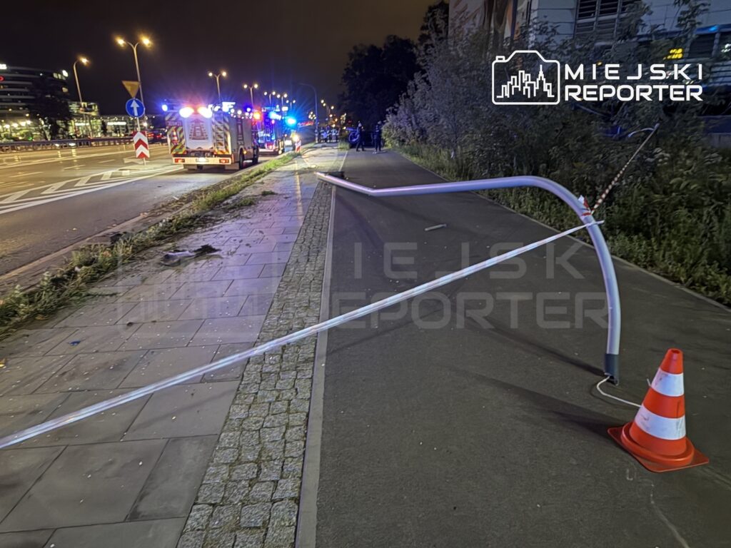 Taśma zabezpieczająca i pachołek odgradzają chodnik przy drodze, gdzie stoją pojazdy służb z włączonymi światłami.