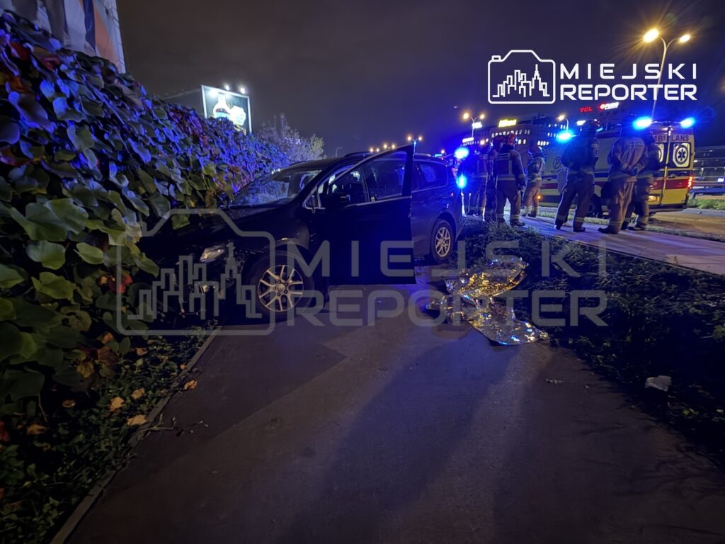 Ciemny samochód stoi na chodniku przy żywopłocie, obok rozbite szkło i służby ratunkowe z karetką nocą.