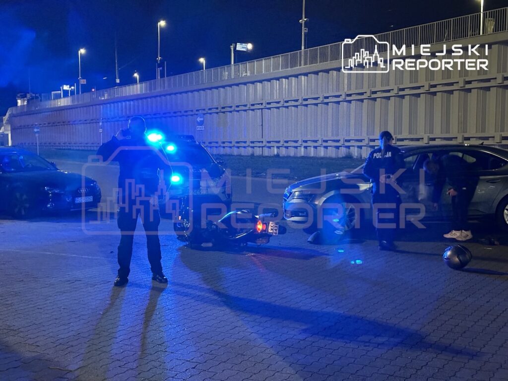 Policjanci przy radiowozie i samochodzie na nocnym parkingu, obok leży kask motocyklowy.
