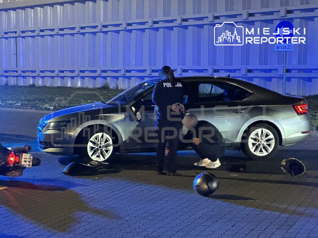 Policjant stoi przy zatrzymanej osobie obok srebrnego samochodu, a na jezdni leżą kaski i elementy motocykla.
