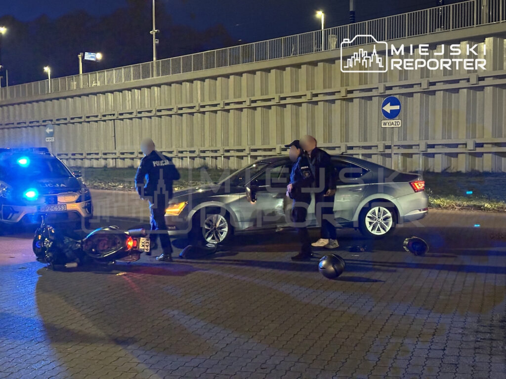 Policjanci stoją przy przewróconym motocyklu i samochodzie po kolizji, nocą pod wiaduktem.