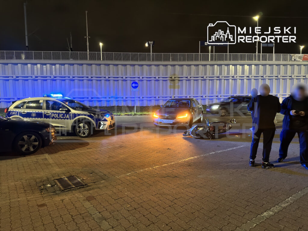Przewrócony motocykl leży na jezdni obok samochodu, radiowozu i dwóch osób nocą.