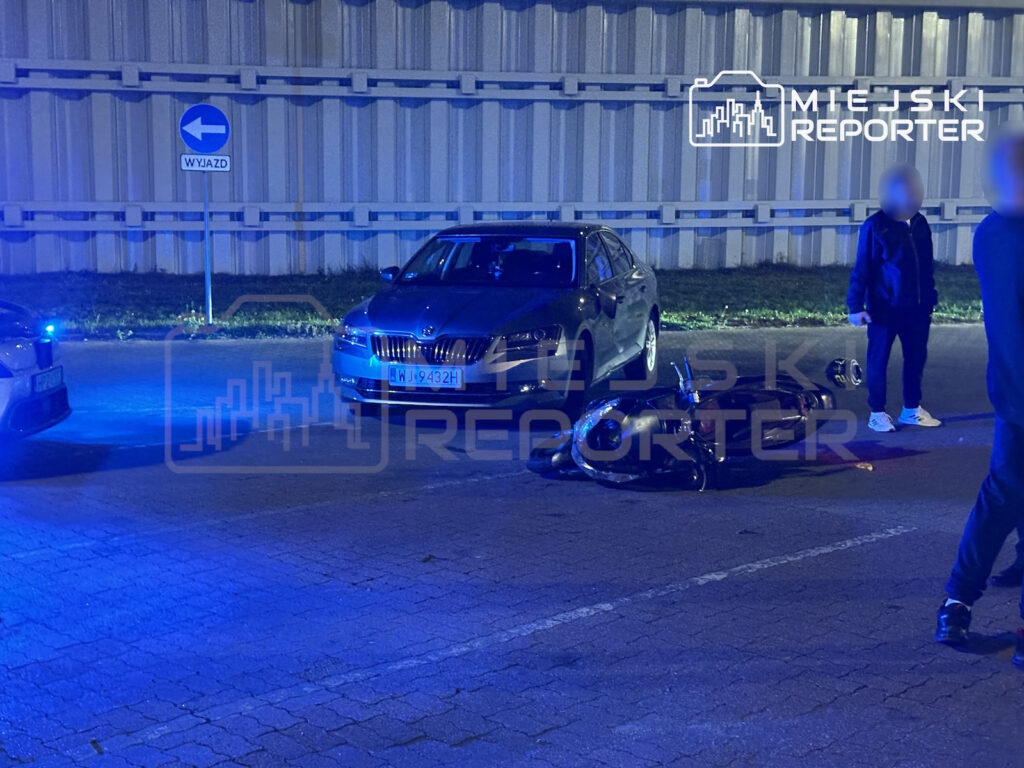 Motocykl leży na jezdni przed samochodem, obok stoją osoby, a miejsce oświetlają niebieskie światła.