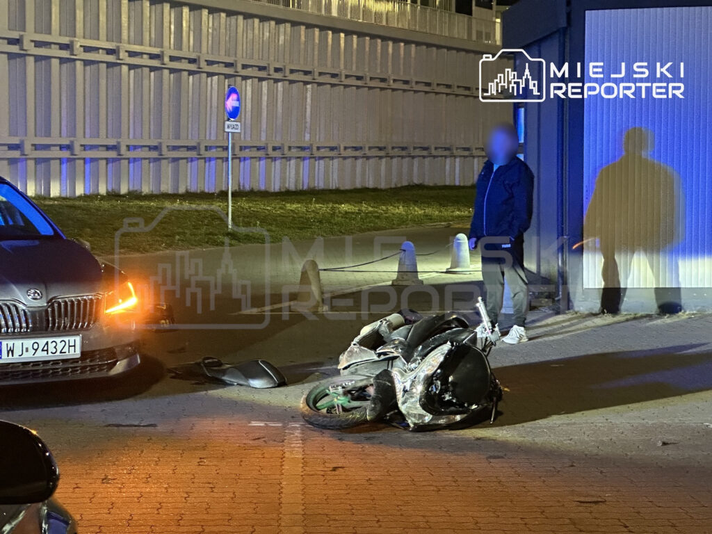 Uszkodzony motocykl leży na jezdni obok samochodu i mężczyzny na chodniku nocą.