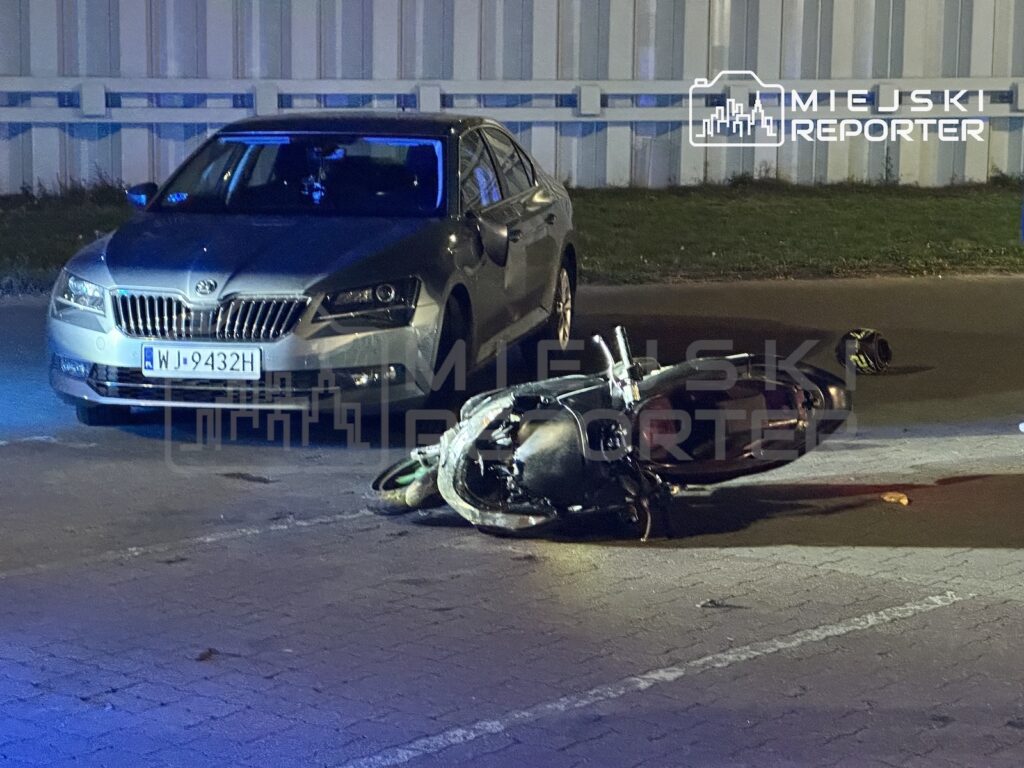Motocykl leży na jezdni przed zaparkowanym samochodem, nocą przy chodniku i budynku.