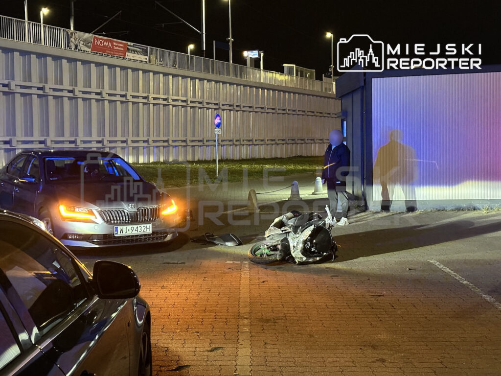 Uszkodzony motocykl leży na jezdni obok samochodu na oświetlonej nocą ulicy.