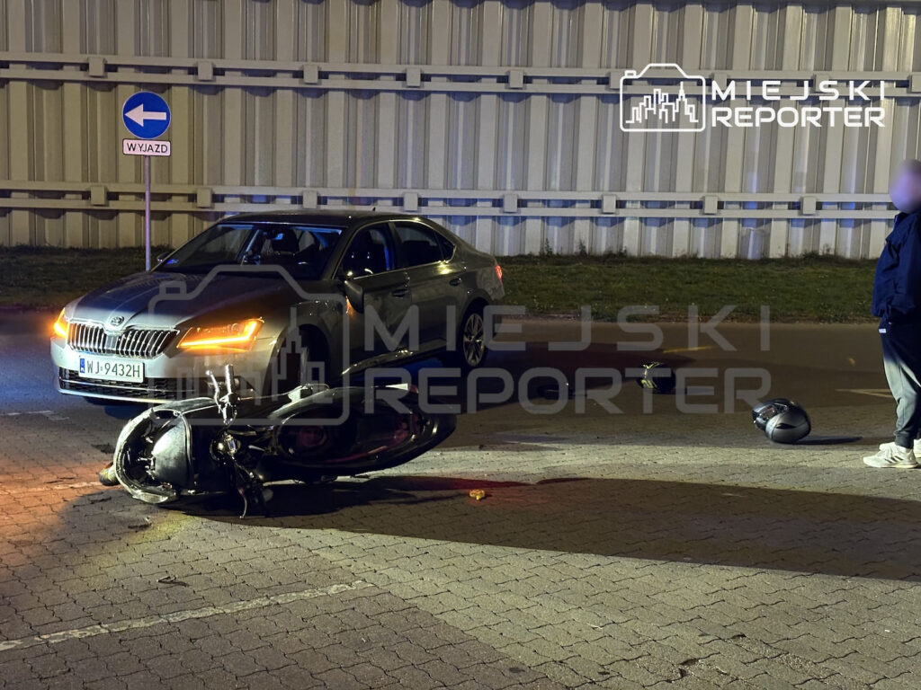 Uszkodzony motocykl leży na jezdni przed samochodem osobowym, obok na drodze leży kask.