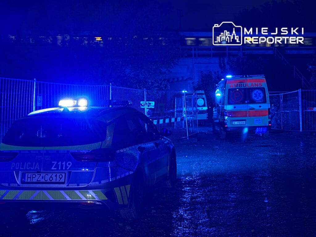 Radiowóz policji i karetka z włączonymi sygnałami świetlnymi przy ogrodzonym terenie nocą.